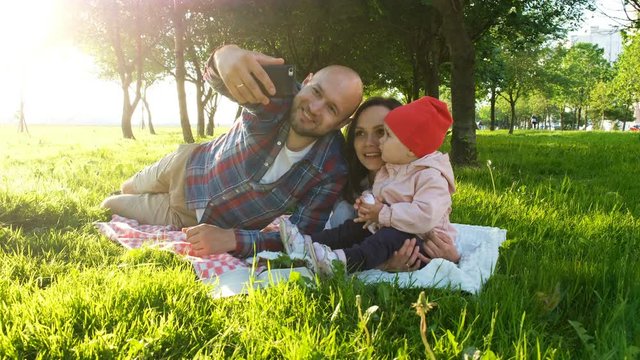 Happy family is laying on the grass and doing selfie with a baby at sunset in the park. Father and mother take pictures of themselves with the baby on the phone