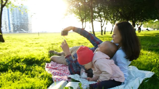 Happy family is laying on the pled and doing selfie with a baby at sunset in the park. Father takes pictures of themselves with the baby