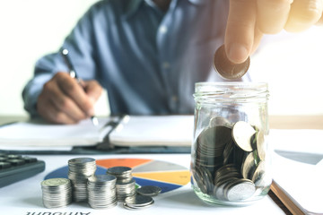 Fototapeta premium Biznes człowiek umieszcza pieniądze lub monetę w szklanym słoju z wykresem i kalkulatorem na biurku do oszczędzania, finansów, koncepcji rachunkowości.