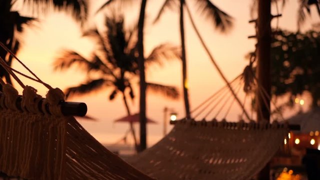 Cozy white hammock on the beach against a background of the swimming pool, ocean and an incredible sunset. Holiday, travel, lounge time concept. Vintage colors and coconut trees at the beach.