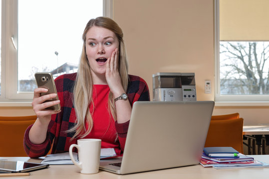 Smiling blonde woman, who is sitting at a laptop and surprised looks at the phone.