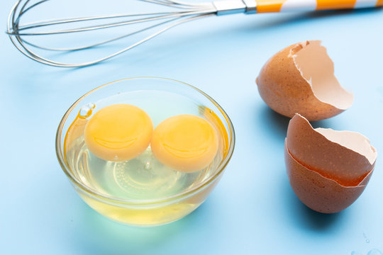 Raw Fresh Egg In Glass Bowl With Eggshell And Whisk On Blue Background