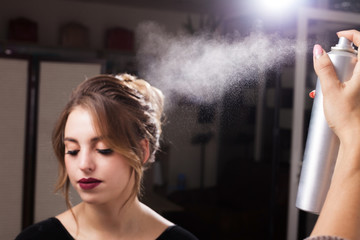 hairdresser fixing a coiffure of a young beautiful model using a hair spray in a beauty salon. girl having a flawless makeup. concept of professional stylist studying