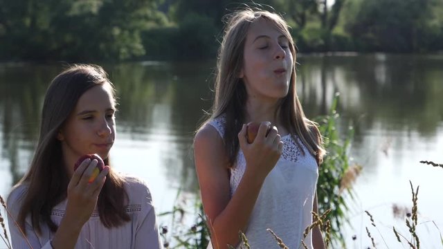 Two sisters teenagers young girls enjoy eat bite apples on nature countryside by a river water