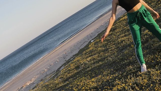 Ragazza ballerina con pantaloni verdi e maglia nera sta ballando allegramente sulla spiaggia in riva al mare