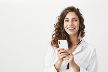 Portrait of happy feminine caucasian woman holding smartphone with both hands, smiling and looking joyfully at camera while standing against gray background. Mom wrote that she reached destination