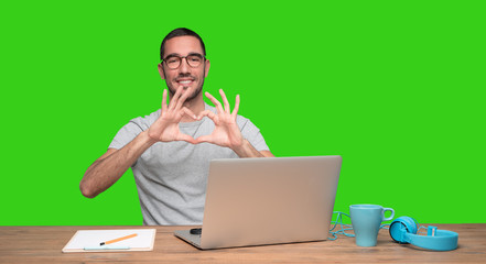 Happy young man doing a gesture of love sitting at his desk - Green background