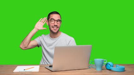 Confident young man trying to listen something with his hand and sitting at his desk - Green background