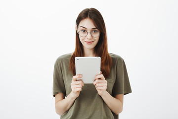 Naklejka premium Good-looking confident brunette girlfriend in trendy round glasses, smiling intriguingly and holding digital tablet, wanting to share with friend new novel she wrote, standing playfully over gray wall
