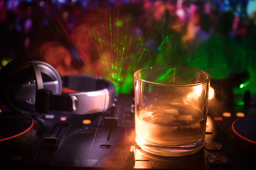 Glass with whisky with ice cube inside on dj controller at nightclub. Dj Console with club drink at music party in nightclub with disco lights.
