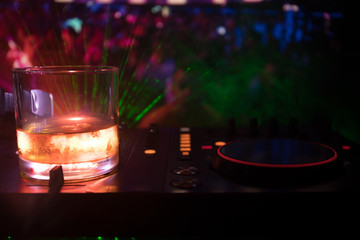 Glass with whisky with ice cube inside on dj controller at nightclub. Dj Console with club drink at music party in nightclub with disco lights.