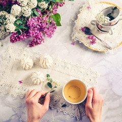 Top view woman's hands holding vintage cup of green tea and spiraea branch on the table with meringue cakes, a bouquet of blossoming lilac and spiraea. Retro style. Selective focus, copy space.