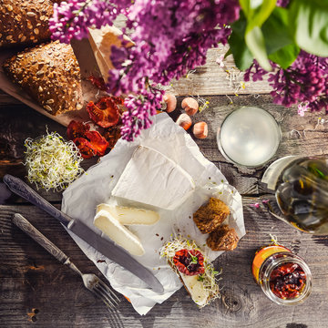 Brie type of cheese. Camembert cheese. Fresh Brie cheese and few slices with bun, dry tomatoes, white wine bottle and glass and lilac on the old wooden rustic table. Italian, French cheese. Top view.