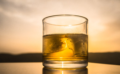 A glass of whiskey with ice on a sunset background or shot of whiskey at sunset dramatic sky on mountain landscape background