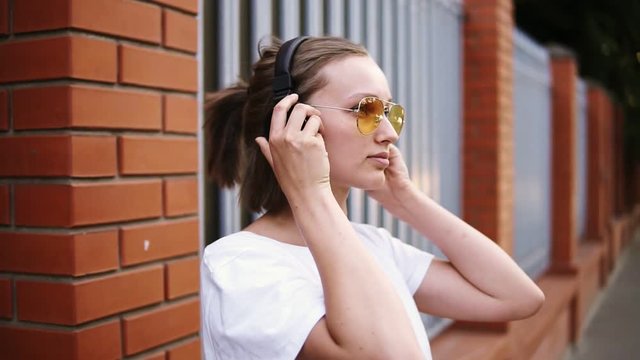 A girl with a short haircut, wearing yellow sunglasses. Relaxed leaning on the fence, listening to her favorite music on black headphones. Enjoy the music. Side view