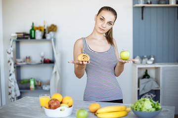 Obraz premium Portrait of a slim attractive woman making a choice between an Apple and a sweet bun, trying to resist temptation
