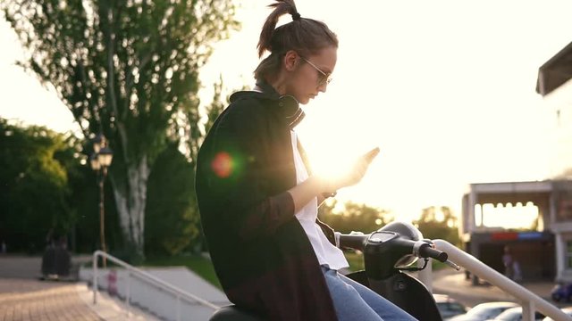Young girl in black hoodie sitting on a motorbike and typing on her mobile. Sitting on moped outside in the park. Sun rays on the background