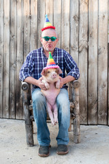 Eccentric elderly farmer with a hat, checkered shirt, blue jeans, glasses and pink rubber gloves plays with funny red piglet in party dunce hat. Hands close-up. Copy space. 2019 Year Yellow Pig