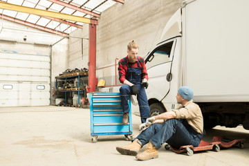 Young bearded male car technician chatting with his colleague while resting near broken truck in large service garage