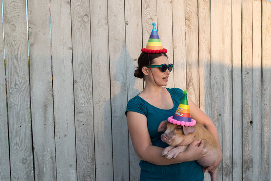 Eccentric slender woman with a hat, green shirtand glasses plays with funny red piglet in party dunce hat. Hands close-up. Copy space. 2019 Year Yellow Pig