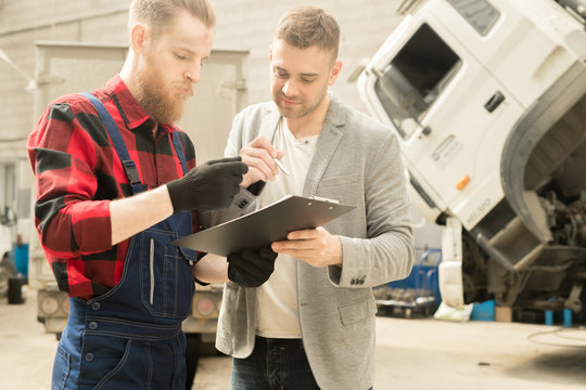 Young bearded auto technician showing inspection checklist to male customer after finished maintenance in repair workshop