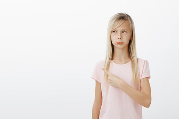 Upset offended cute european girl with blond hair in trendy pink shirt, pointing at upper left...