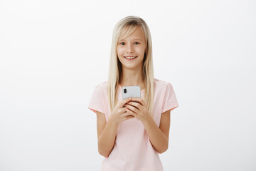 Adorable positive child feeling great finally talk with father who works abroad. Satisfied cheerful cute blond girl in pink t-shirt, holding expensive smartphone and smiling broadly at camera