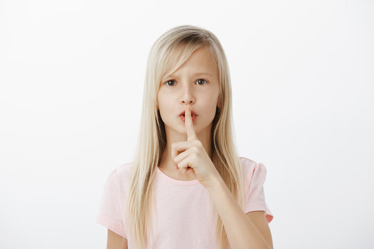Studio shot of sctrict confident little blond girl in casual pink t-shirt, saying shh while showing shush gesture with index finger over mouth, asking to be quiet while looking after baby brother