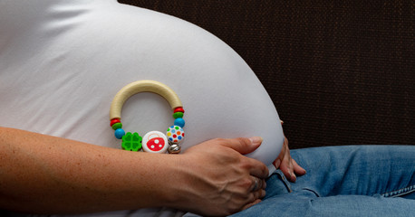 Pregnant woman with baby tummy sitting on the sofa holding her baby belly and lying a baby toy her her tummy