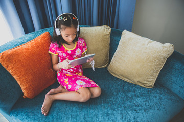 Little girl sitting on sofa listening music from headphone computer tablet