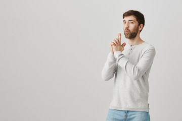 Mature man plays with kids and acts like soldier. Studio shot of childish father showing weapon or gun gesture, blowing at hand as if making shot, standing over gray background with serious face