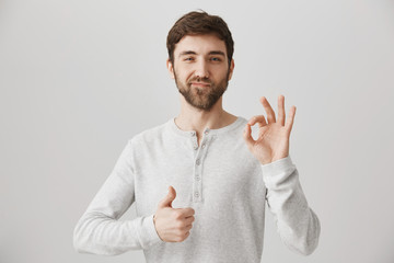 Handsome and confident european guy with beard showing okay or fine gesture and thumbs up, expressing pleasure or satisfaction while being agree with speaker, standing over gray background.