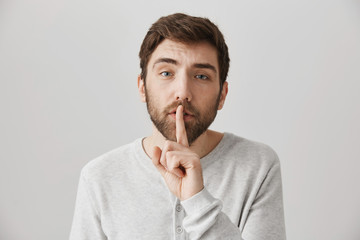 Close-up portrait of confident tired male with beard making shush gesture while standing over gray background. Man asks friends to keep voice down cause he suffers hangover. Secret concept