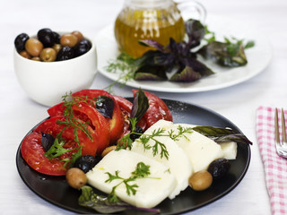 White cheese with tomatoes, green and black olives, basil, coriander and olive oil on a black plate