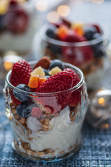 A healthy colourful snack consisting of granola and yoghurt with assorted berried and dried fruits in a small glass cup.

