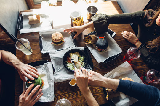 Friendship, Holidays, Fast Food And Celebration Concept - Cropped Human Hands Grabbing Drinks And Snacks Eating Burgers At Diner