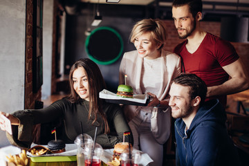Group of positive cheerful friends having fun, taking selfie with smartphone in burger bar. Young blonde woman holding tray with ready green burger