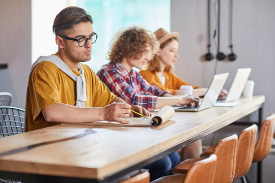Three young co-workers sitting by table and carrying out individual work in office