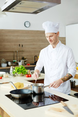 Young chef standing by electric stove and mixing food in pan during process of cooking