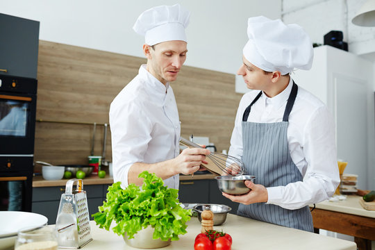 Chef with whisk mixing raw eggs in steel bowl while consulting his trainee in the kitchen