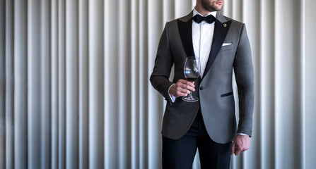 Man in custom tailored tuxedo, suit holding glass with wine and posing indoors in front of...