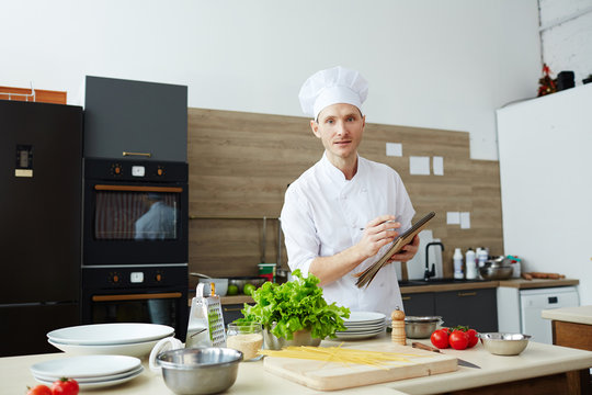 Young chef in uniform making working notes in notepad in the kitchen during process of cooking