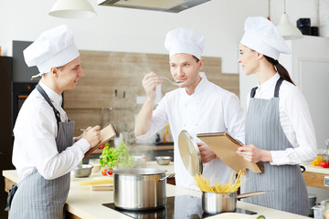 Chef tasting hot food from pan while his trainees making notes near by