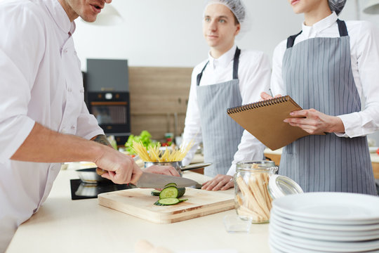 Woman in apron making cooking notes in notepad while chef slicing cucumber on wooden board