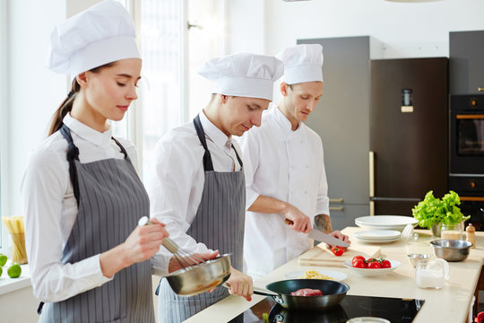 Young woman mixing ingredients in bowl while her colleague frying meat near by and chef preparing salad
