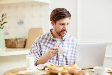 Young businessman with cup of tea sitting in the kitchen with laptop in front and reading online news