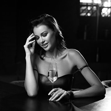 Elegant young woman sitting in bar and having good time alone. Pretty brunette girl in black leather dress. Lady drinking glass of wine.