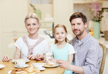 Young man, woman and their daughter looking at camera during breakfast in the kitchen