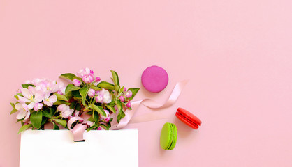 Flat lay top view White gift bag with spring flowers and cake macaron or macaroon on pink background, beautiful dessert, colorful almond cookies, pastel colors