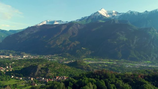 Aerial: flying over valley and green forest in springtime, snowcapped mountain range and villages, Susa Valley, the Alps, Turin, Italy.